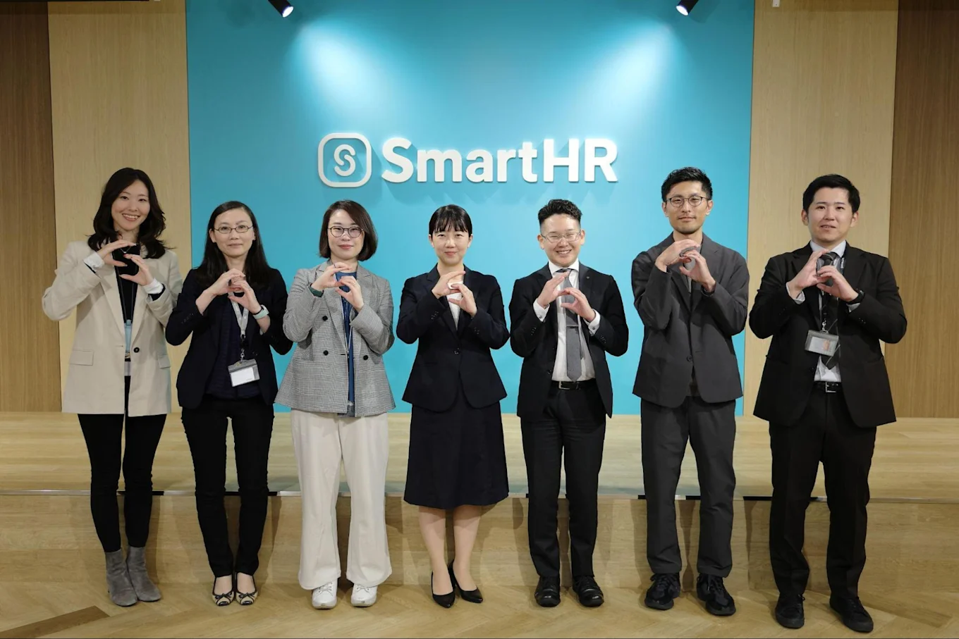 SmartHRのロゴが飾られた青い壁の前に鵜澤さん、近藤さん、ほかSmartHR従業員など。皆SmartHRロゴマークのハンドサインをしている