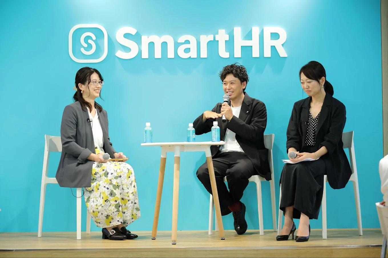 SmartHRのロゴが掲げられた青い背景のステージで、マイクを持ち座って対談する3人。左から松栄、須藤さん、渋谷さん。