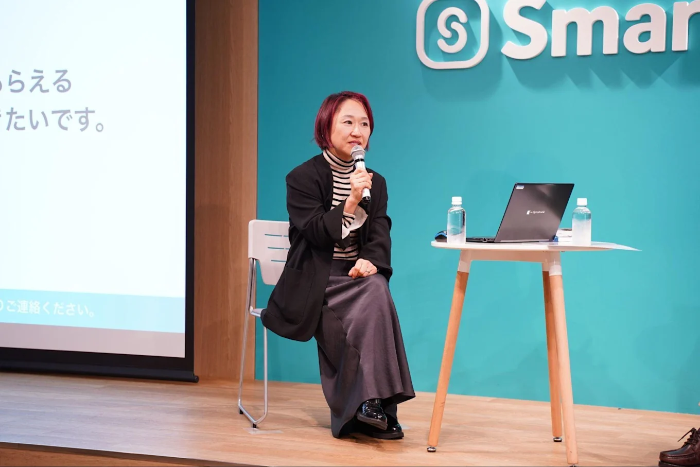 SmartHRのロゴがあしらわれた壁の前で話す山崎さん。マイクを片手に椅子に座っている
