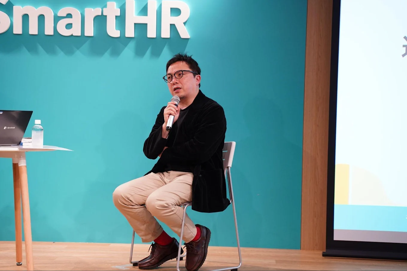 SmartHRのロゴがあしらわれた壁の前で話す古賀さん。片手にマイクを持ち、もう片腕を反対の腰に当てるように組み、椅子に座っている
