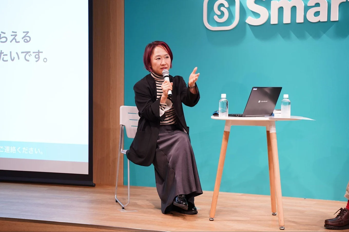 SmartHRのロゴがあしらわれた壁の前で身振りを加えて話す山崎さん。片手にマイクを持ち椅子に座っている