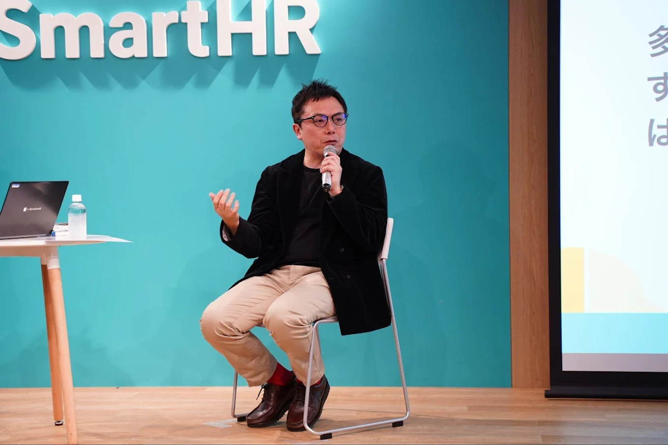 SmartHRのロゴがあしらわれた壁の前で話す古賀さん。マイクを片手に椅子に座っている