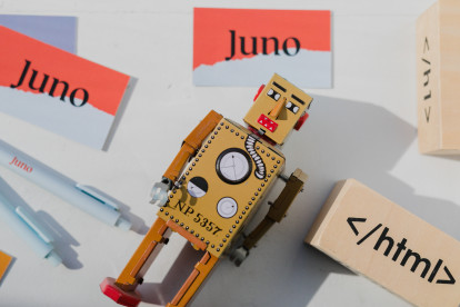 Toy robot lying on a table with a wooden building block with a HTML tag on it, and Juno-branded business cards and pens.