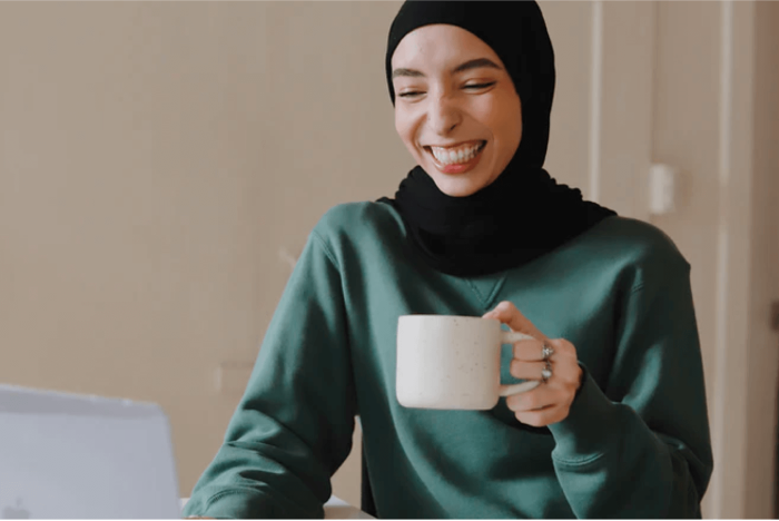 Juno College student wearing a black hijab and holding a cup of tea, smiling at a laptop camera