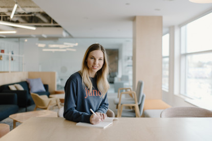 Heather Payne in a brightly-lit common space at Juno's campus. She's smiling at the camera while writing in a notebook, and wearing a Juno sweatshirt.