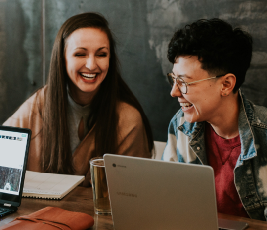 Two Juno College students are sitting at a table with their laptops and laughing