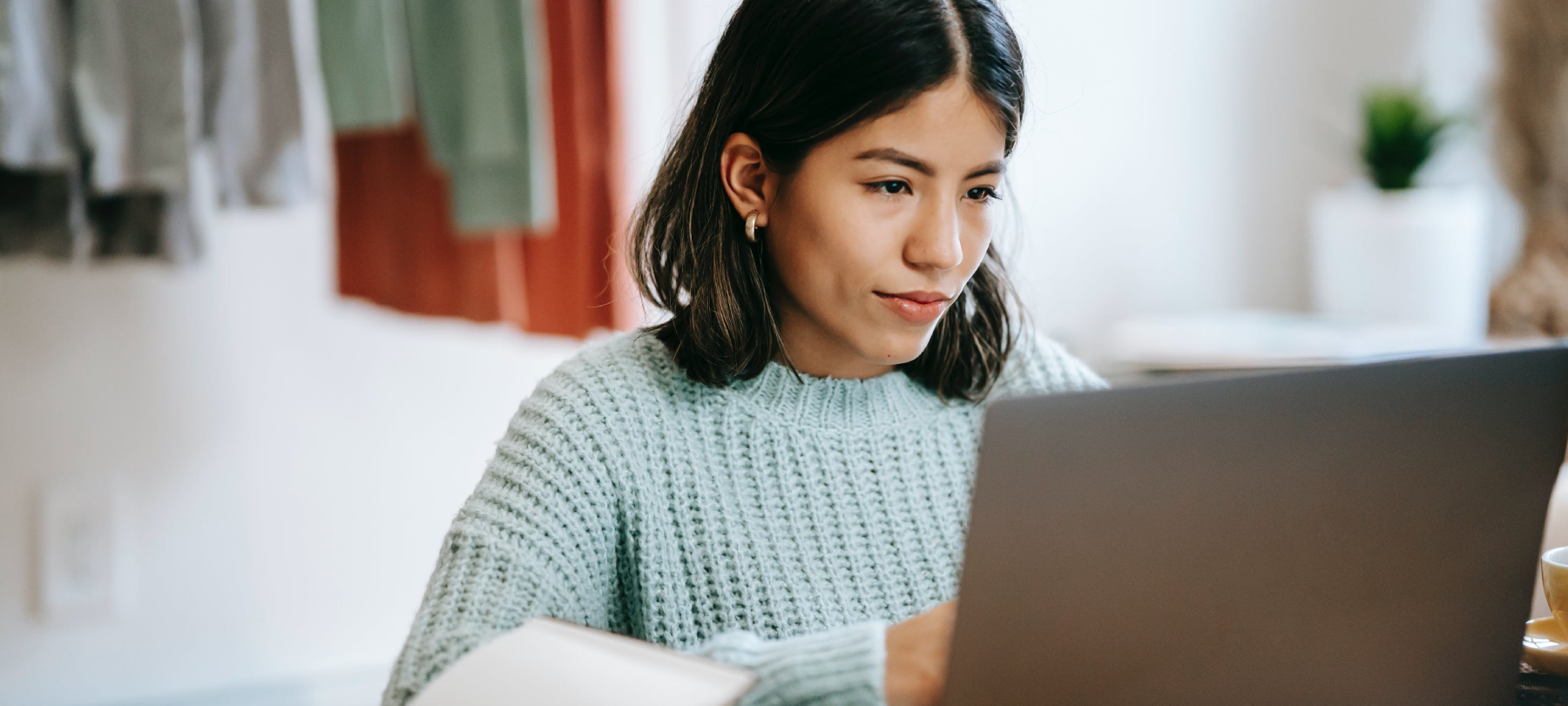 A woman in a knit sweater types on her laptop in her bedroom