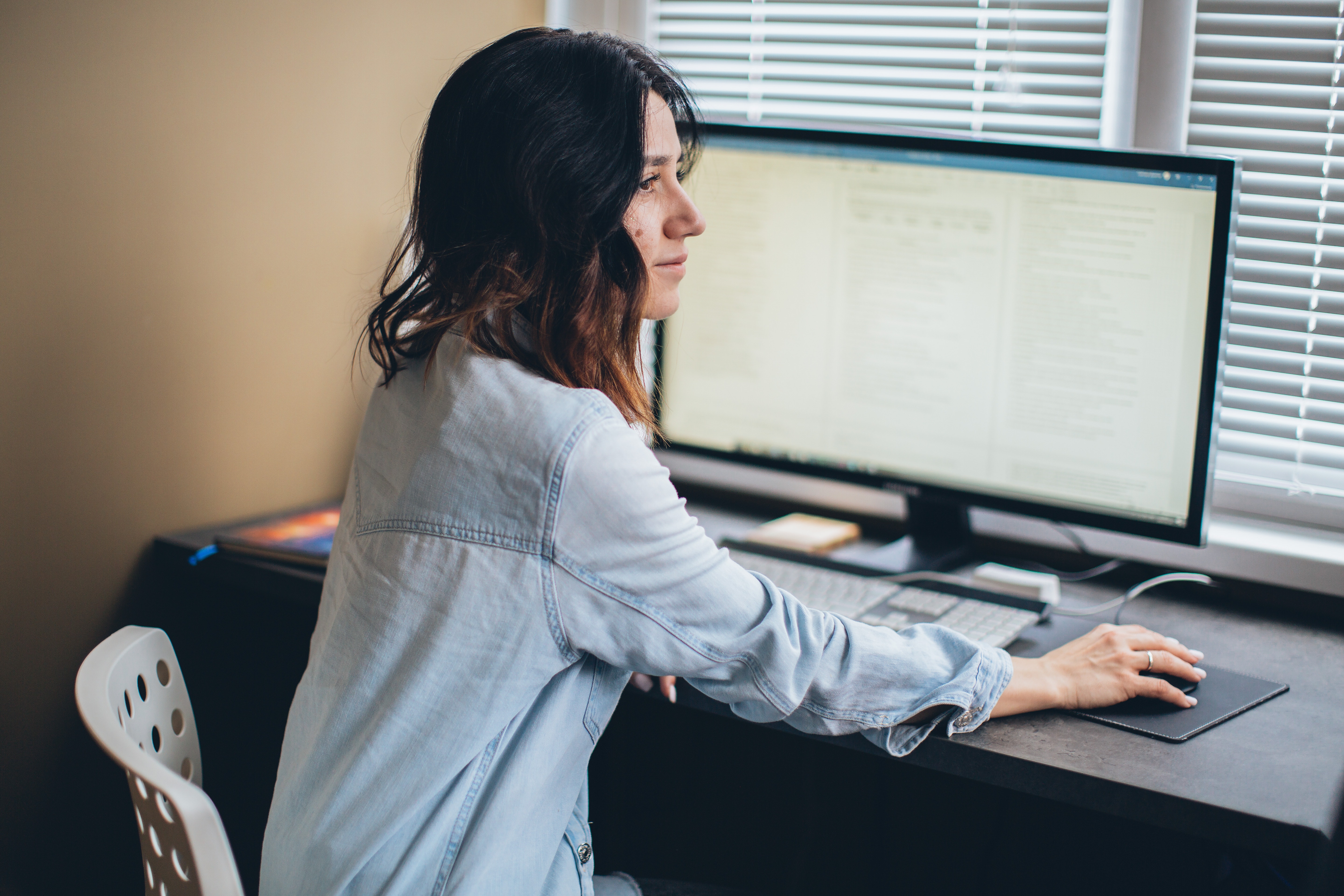 A woman sits in front of her desktop computer, looking pensively into the distance.