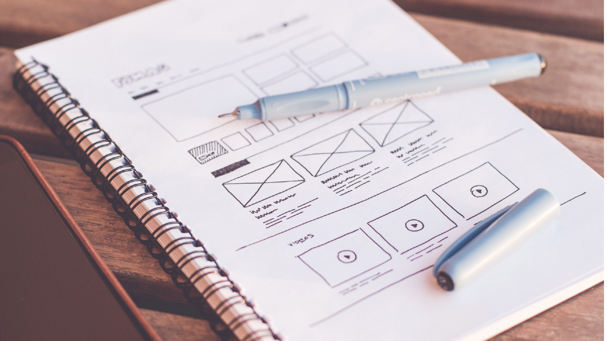 A notebook with a wireframe drawing of a website/app in pen. A pen lies on the notebook, uncapped.