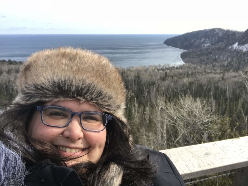 Jessica Duarte outside, smiling and wearing a furry hat and a winter coat. The backdrop is a lake and forest.