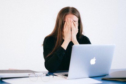 Woman in front of her laptop on a desk with her hands covering her face in stress