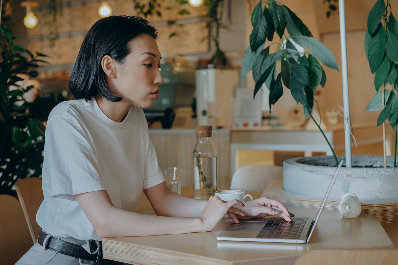 A woman typing on her laptop in a cafe, surrounded by potted plants.
