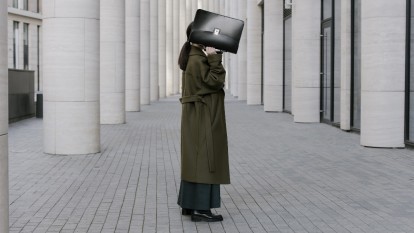 Person in business professional clothes holding up a briefcase, covering their face. They're standing on concrete, surrounded by concrete pillars.