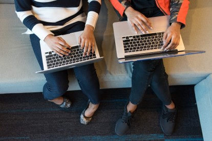Overhead shot of two women sitting on a couch typing on their laptops