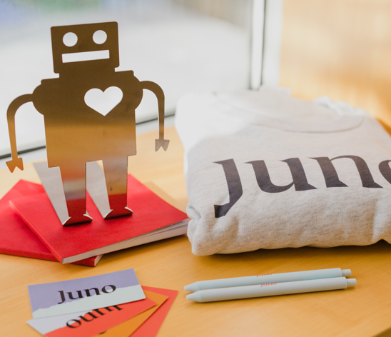A paper cut out of a robot with a square head and big heart stands next to Juno merchandise that includes a sweater, pens, and cards.