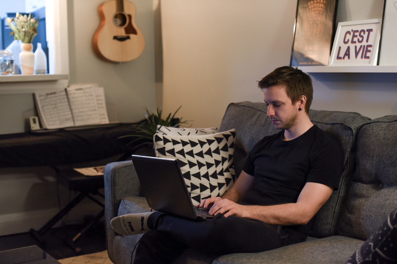 Student working at their laptop on a grey couch in a room with a guitar on the wall behind him.
