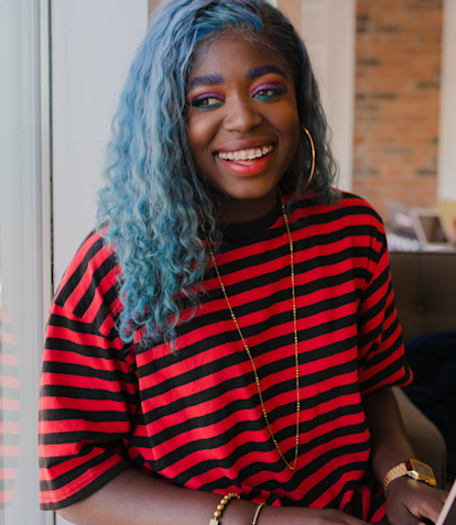 Woman with dyed blue hair smiling at the camera and sitting on a couch at Juno College, with a laptop on her lap.