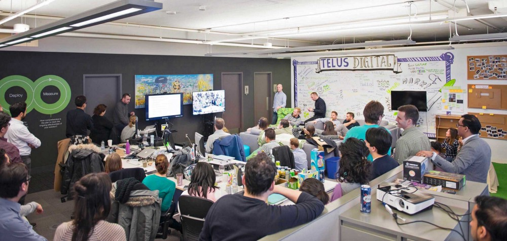 A large group of people sit in an office, listening to someone present using slides, a giant whiteboard labeled TELUS Digital along one wall