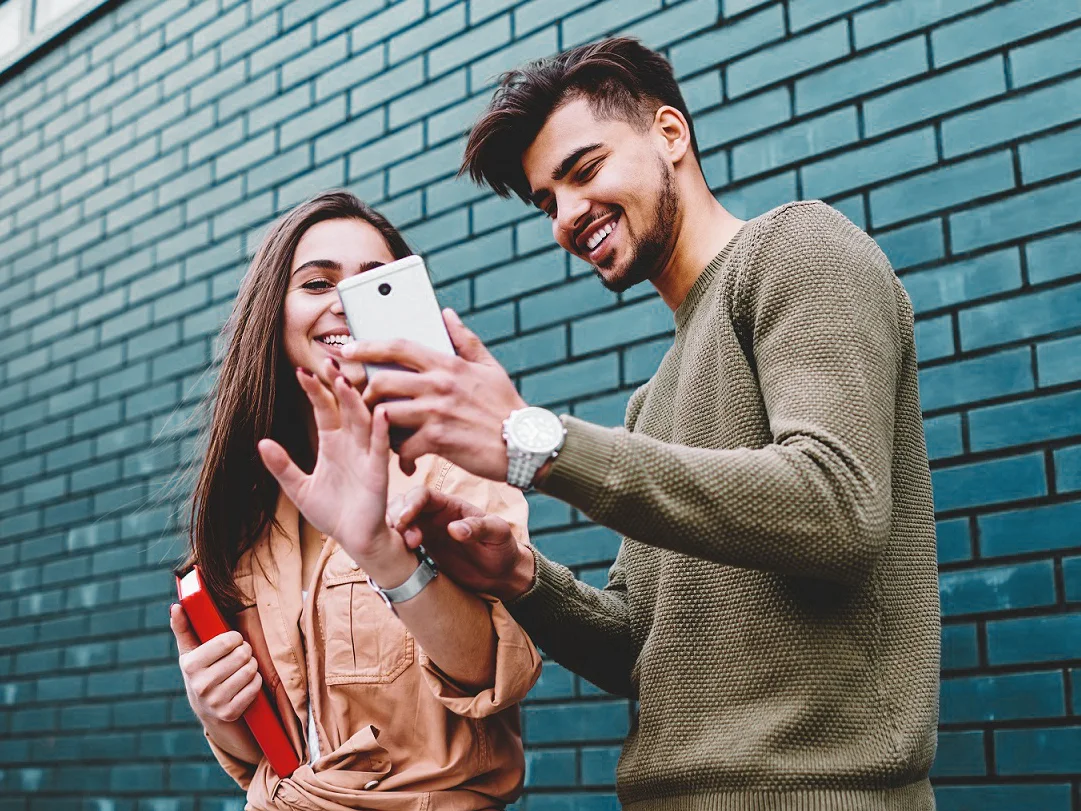 Un jeune homme et une jeune femme sourient en regardant l’écran d’un téléphone intelligent