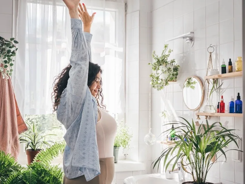 Woman in a bright, plant-filled bathroom stretching her arm upward in a relaxed morning wellness pose, with natural light streaming through the window and organized shelves of skincare products visible in the background."