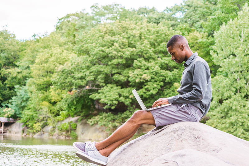 A man sitting on a rock in nature working on a laptop.