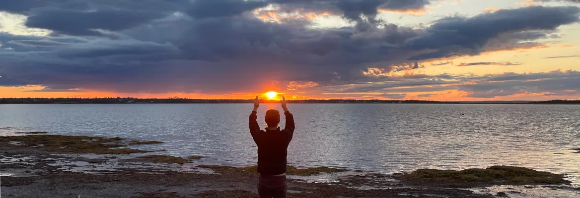 A person stands on a beach with the sun setting on the horizon. Their hands are uplifting as if cradling the setting sun between their hands.
