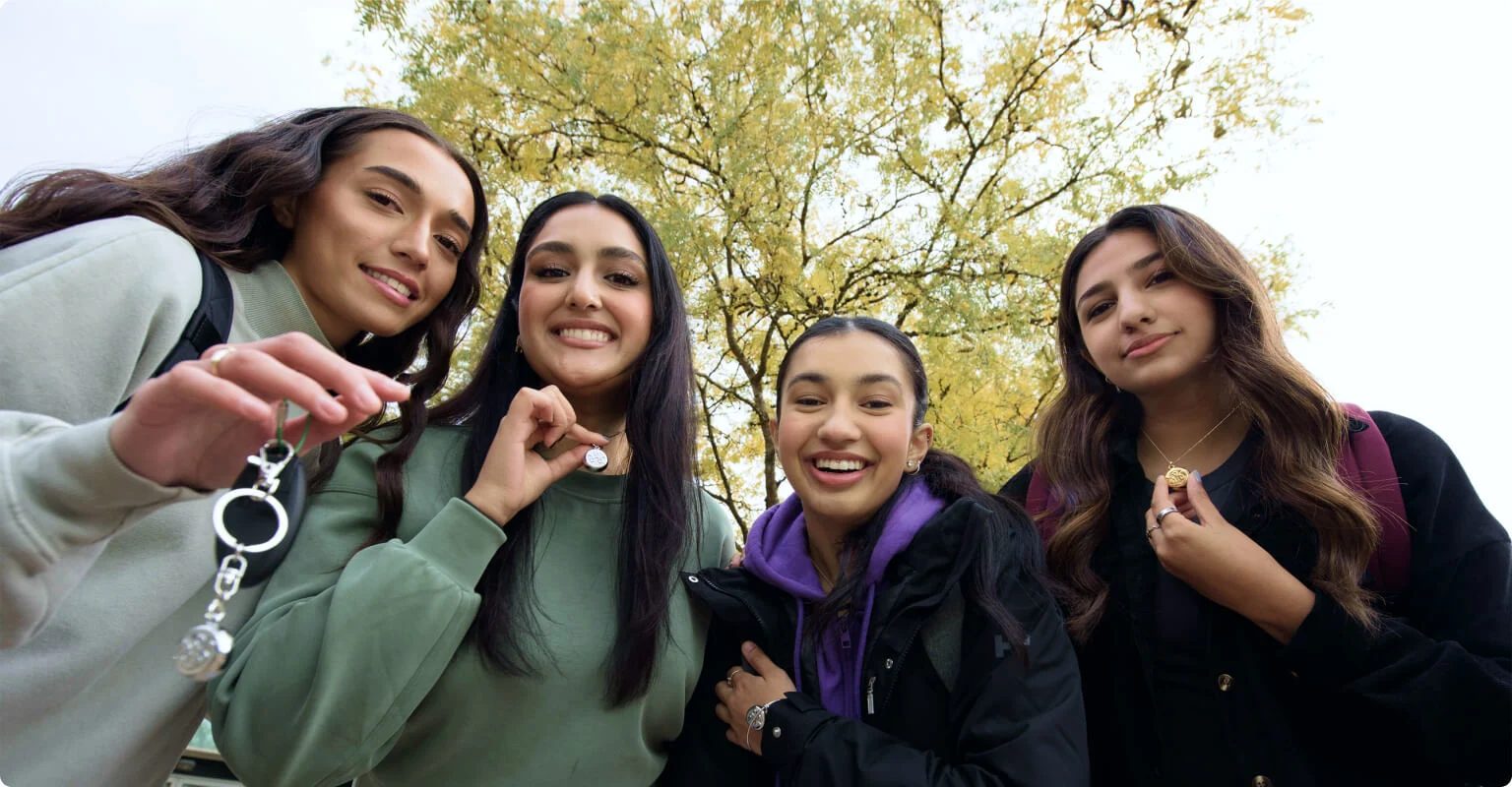 Un groupe de jeunes gens regardant la caméra, souriant et montrant leurs Accessoires de sécurité connectés de TELUS sous forme de porte-clés, de colliers et de bracelets, de couleur or et argent.