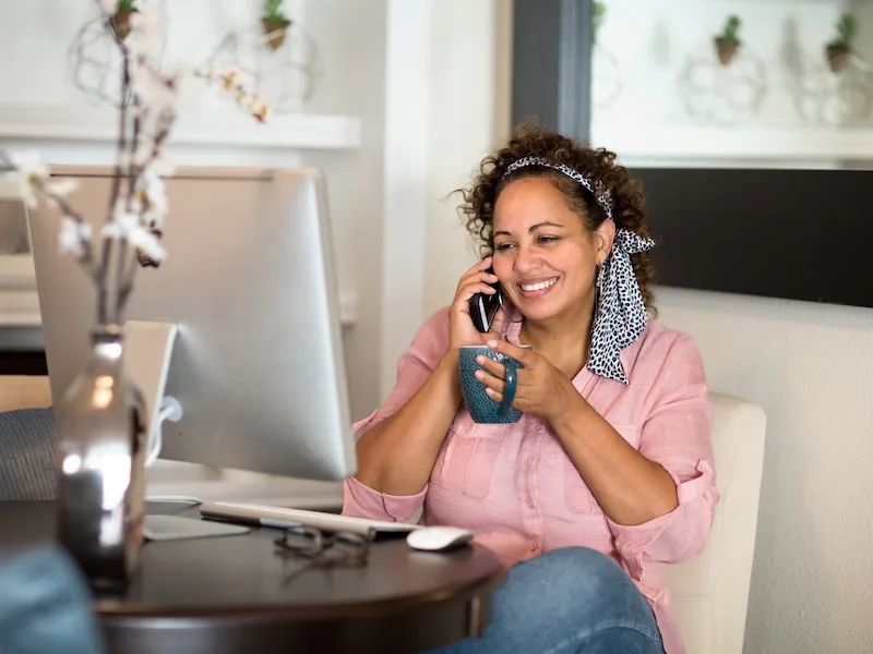 Happy woman in a pink shirt working from home, holding a cup of coffee while having a phone conversation at her desk with a computer monitor