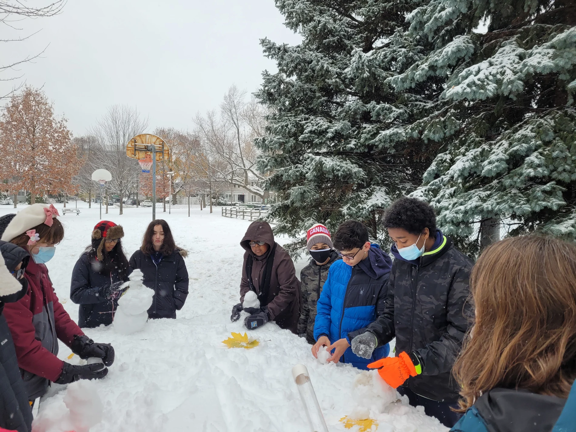 A group of students in the schoolyard, gathered around and playing in the snow. 
