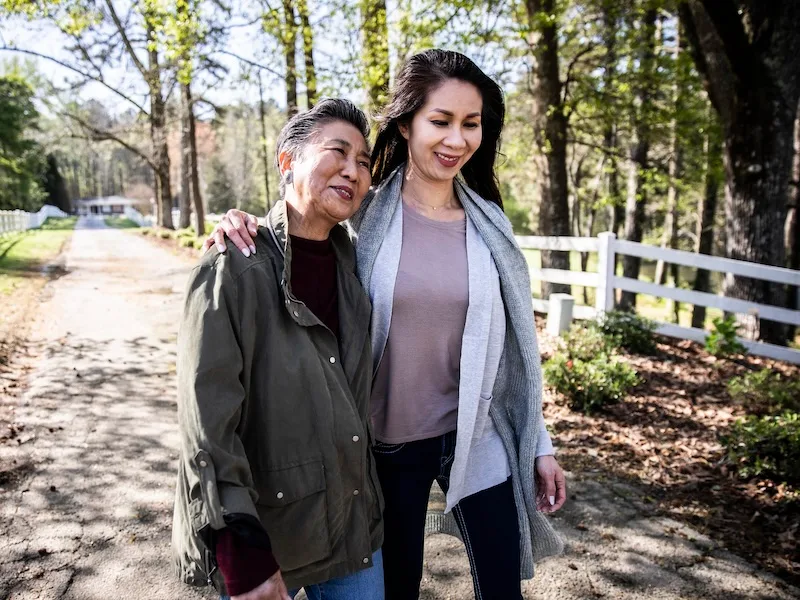 An older man and younger woman standing together smiling outdoors on a rural road with trees and fence in the background.