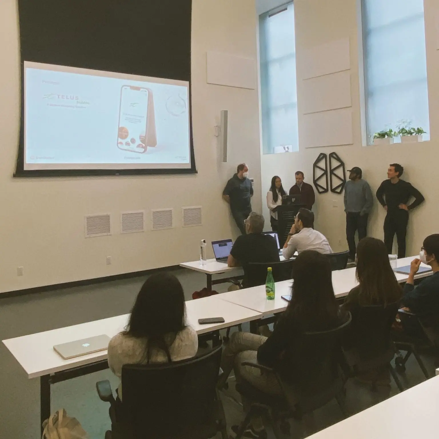 5 people stand at the front of a classroom, presenting work on a screen while people sitting at long white desks look on