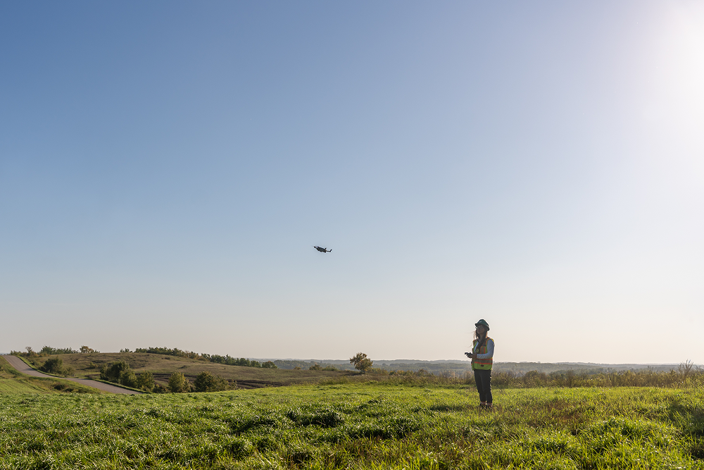 Field worker standing in a grassy field operating a flying drone against a clear blue sky.