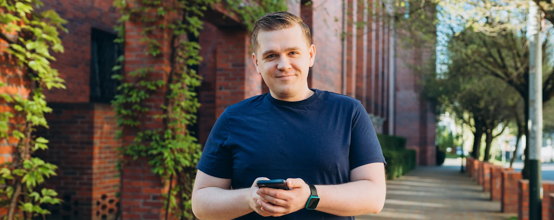 Man in a navy blue t-shirt holding a smartphone while standing on a brick pathway surrounded by ivy-covered brick buildings and trees in an urban setting.