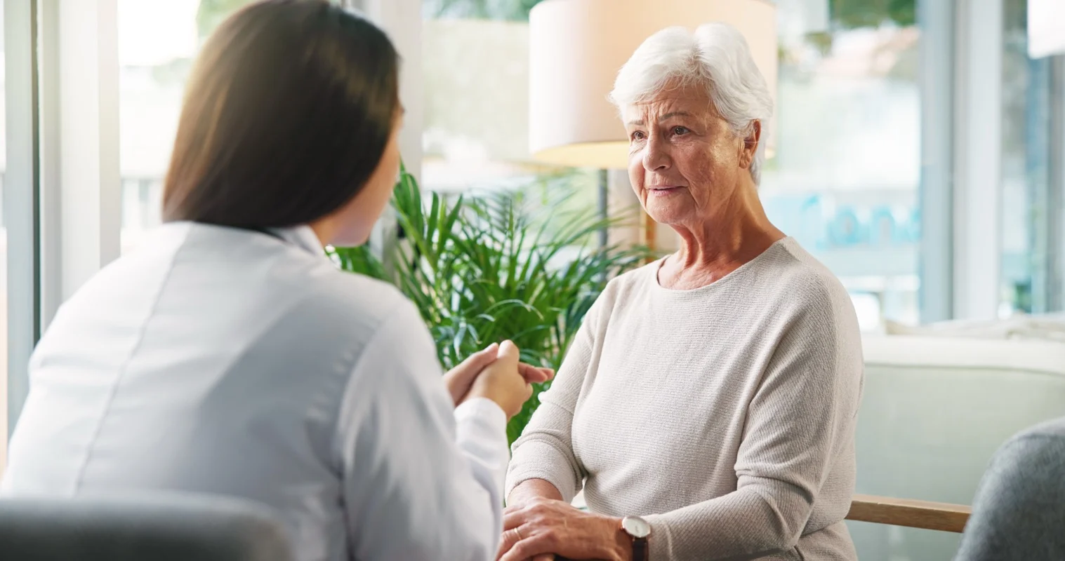 A healthcare professional in a white coat holds an older woman's hand during a conversation in a home setting