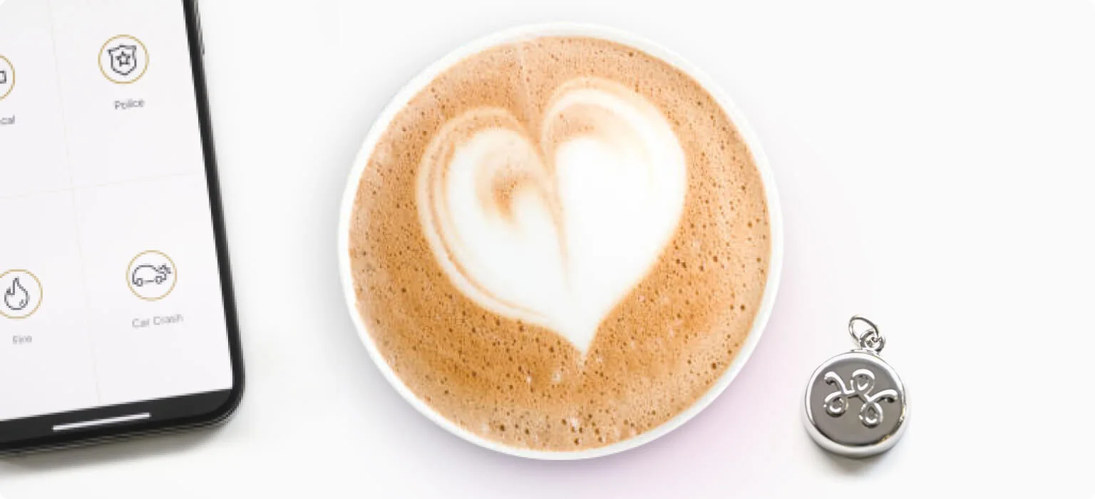 A smart phone, sitting next to a cup of coffee with a heart drawn in the coffee’s foam topping, sitting next to a TELUS SmartWear security charm in silver color.
