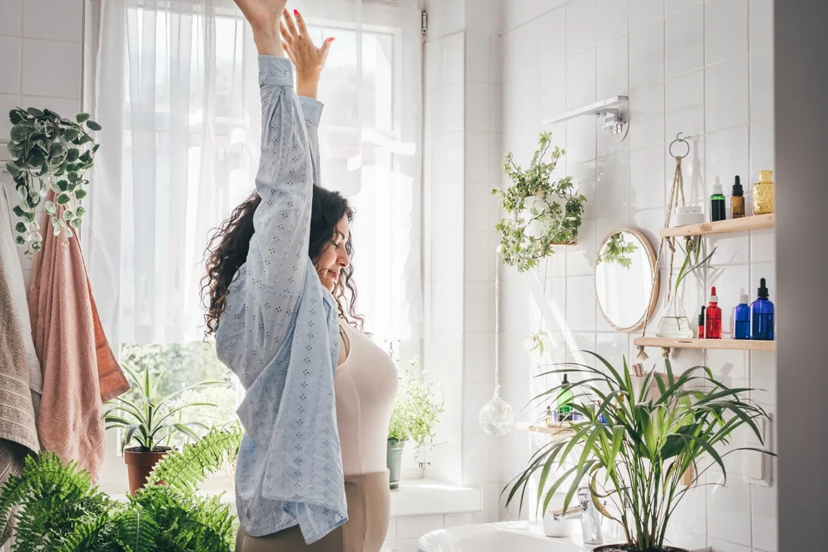 Woman in a bright, plant-filled bathroom stretching her arm upward in a relaxed morning wellness pose, with natural light streaming through the window and organized shelves of skincare products visible in the background.