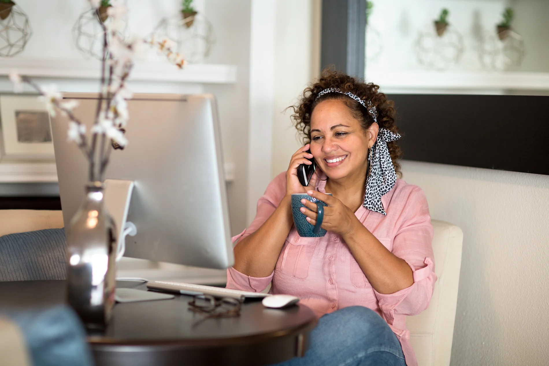 Happy woman in a pink shirt working from home, holding a cup of coffee while having a phone conversation at her desk with a computer monitor