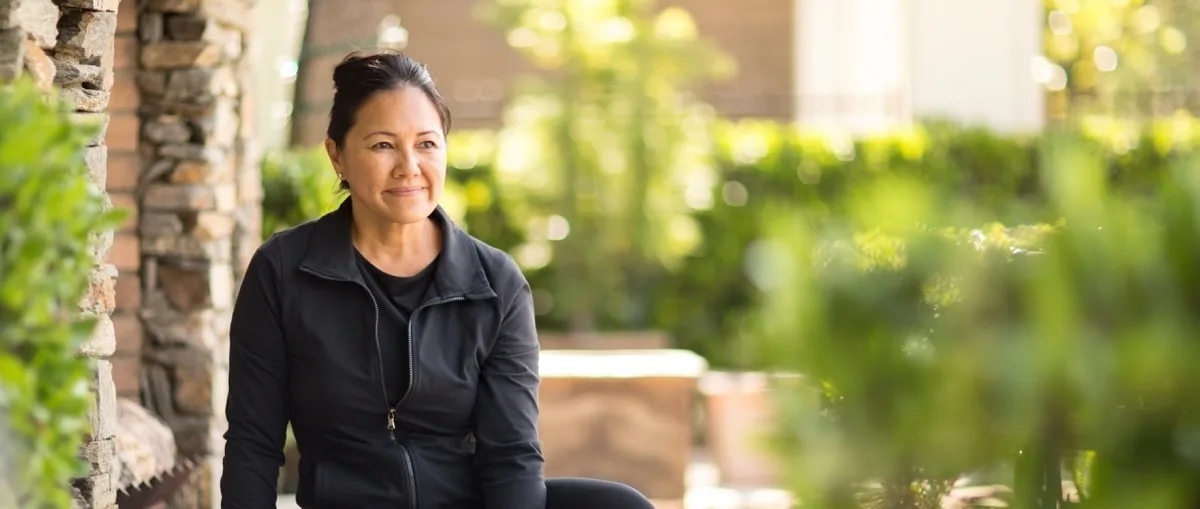 Middle-aged woman in black athletic wear sitting on a stone bench outdoors, smiling.