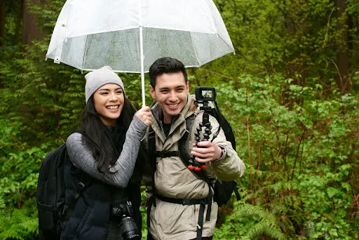 Two young adults are enjoying the outdoors while standing under an umbrella and smiling in front of a camera.