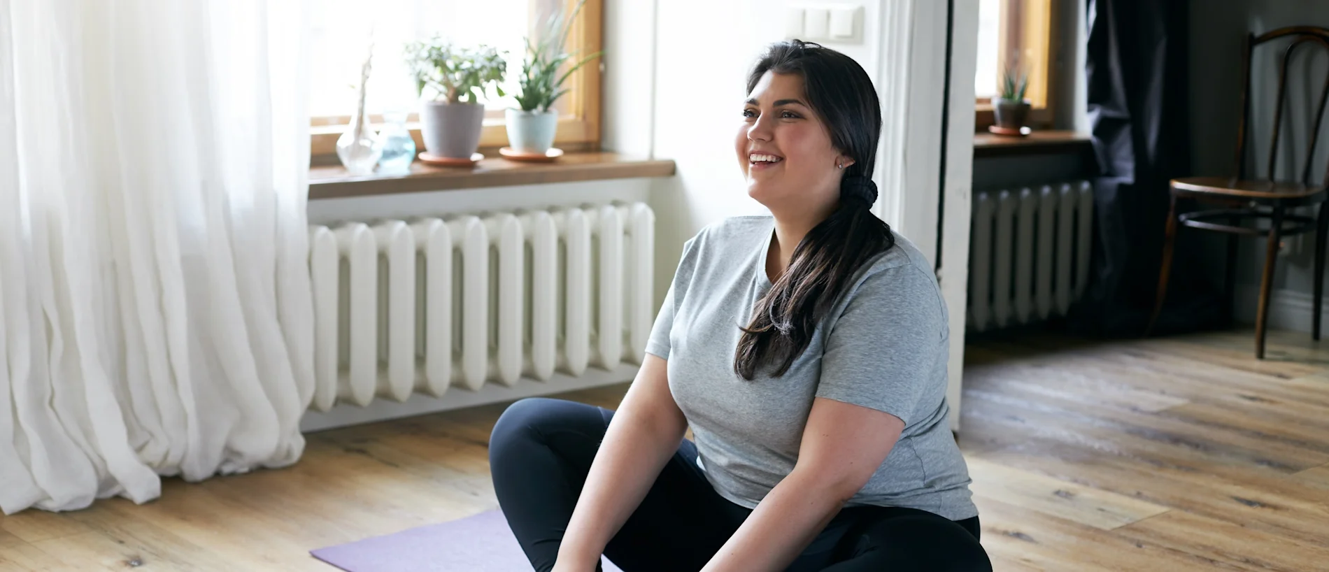 Smiling woman in gray shirt sitting cross-legged on a purple yoga mat in a bright bedroom, surrounded by plants and natural light