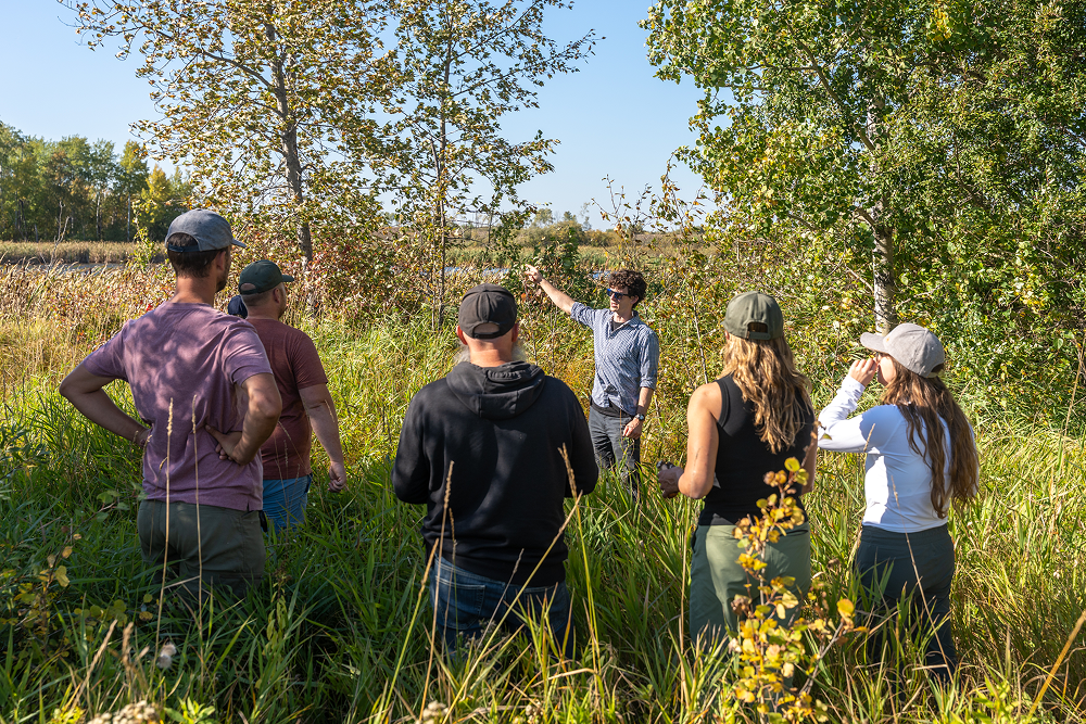 A man points toward trees while speaking to a group standing in tall grass outdoors on a clear day.