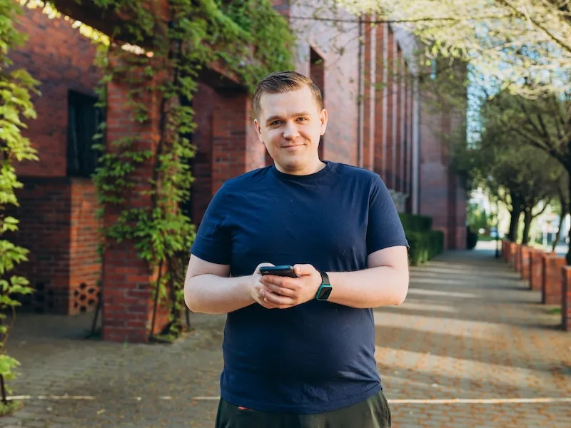 Man in a navy blue t-shirt holding a smartphone while standing on a brick pathway surrounded by ivy-covered brick buildings and trees in an urban setting.