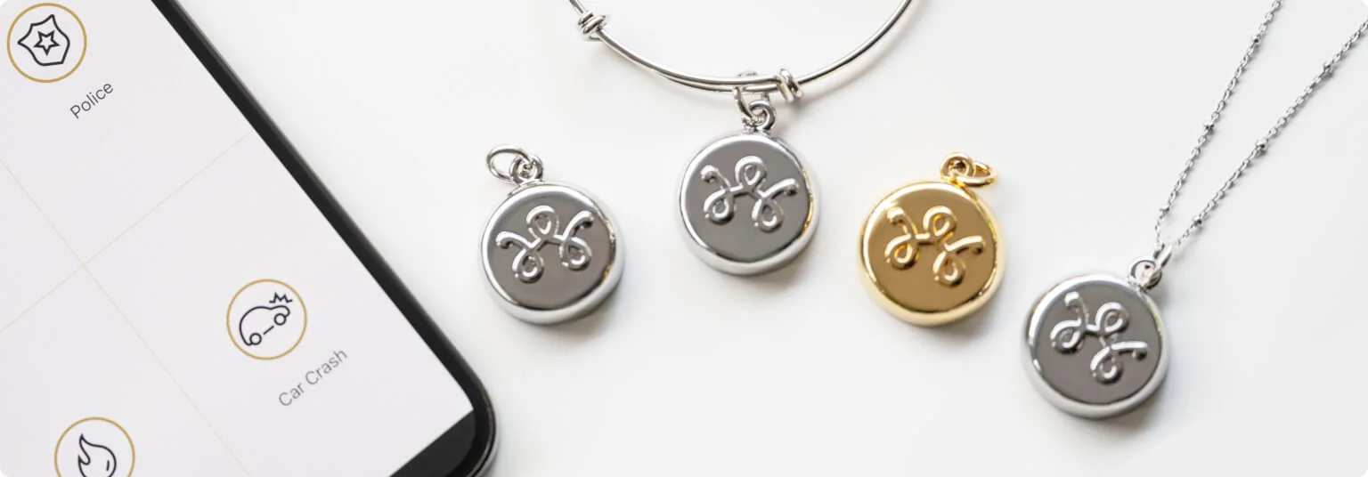 A smart phone sitting next to a collection of TELUS SmartWear security devices, including the silver-colored keychain charm, the silver-colored bracelet, the gold-colored charm, and the silver-colored necklace.