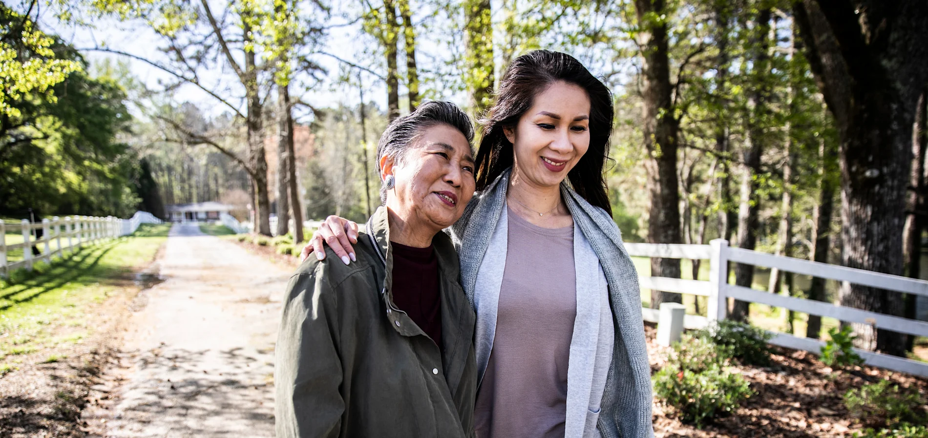 An older man and younger woman standing together smiling outdoors on a rural road with trees and fence in the background.