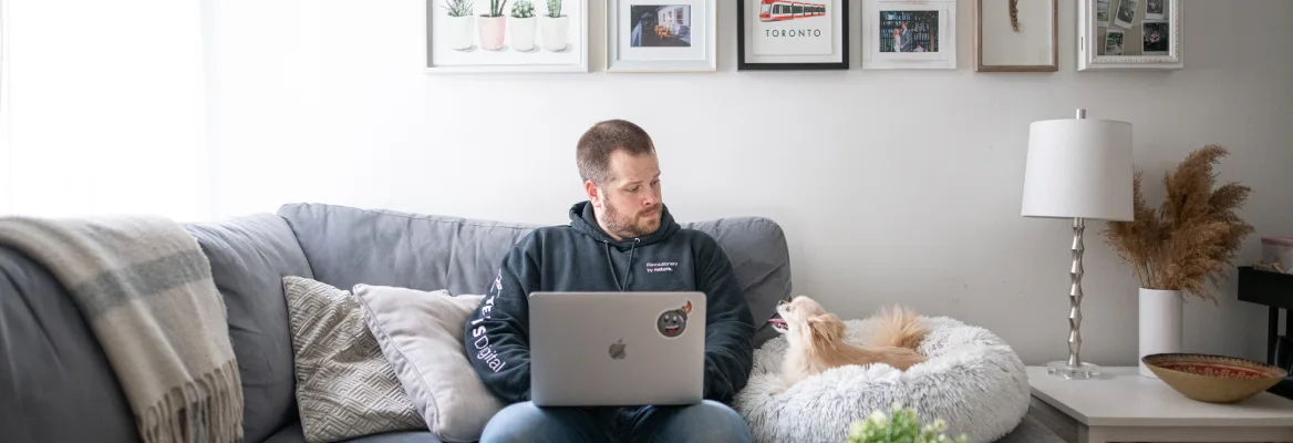 A bearded man in a hoodie sits on a sofa on a laptop, small dog next to him in a round dog bed