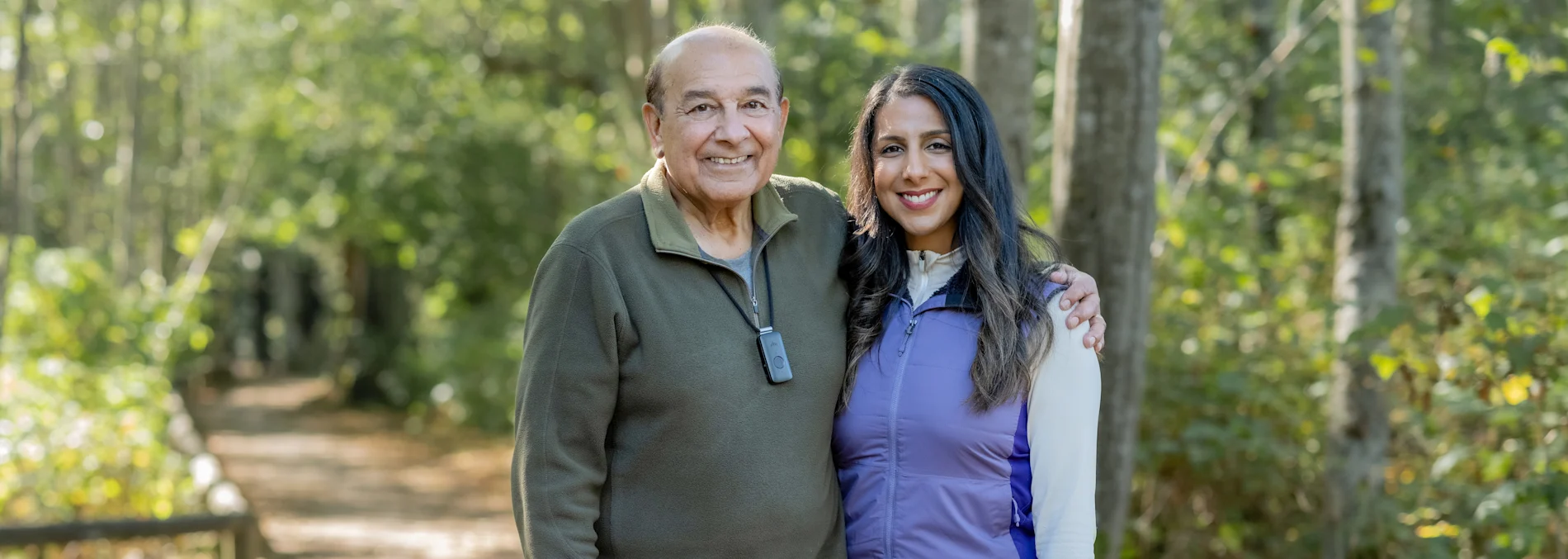 An elderly man and his daughter smile together on a wooded trail. He wears a discreet emergency pendant, demonstrating how medical alert systems for seniors Canada provide peace of mind and safety for active families enjoying the outdoors.