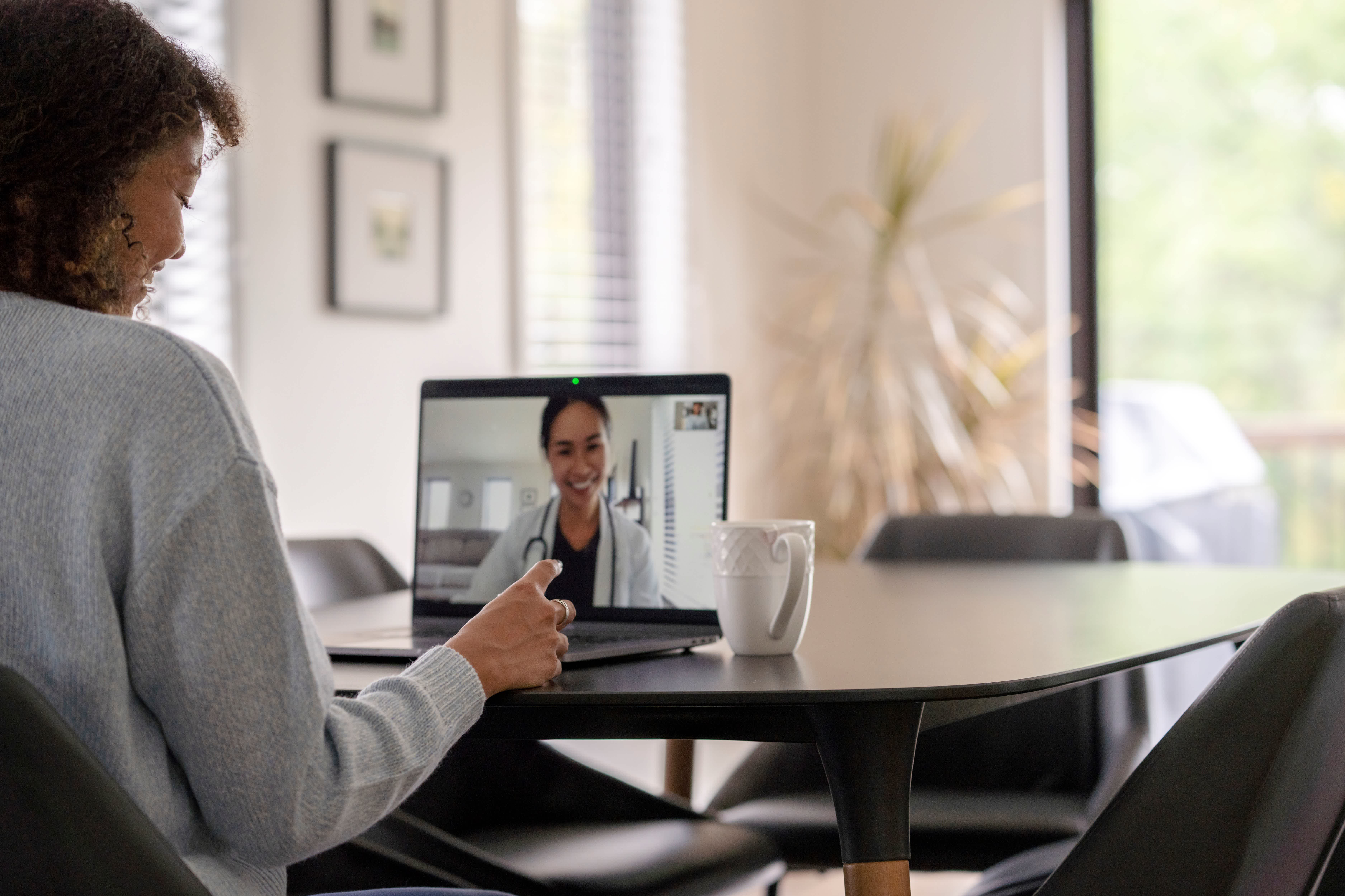 Woman sitting at a dining room table having a virtual consultation on her laptop with a female nurse practitioner.