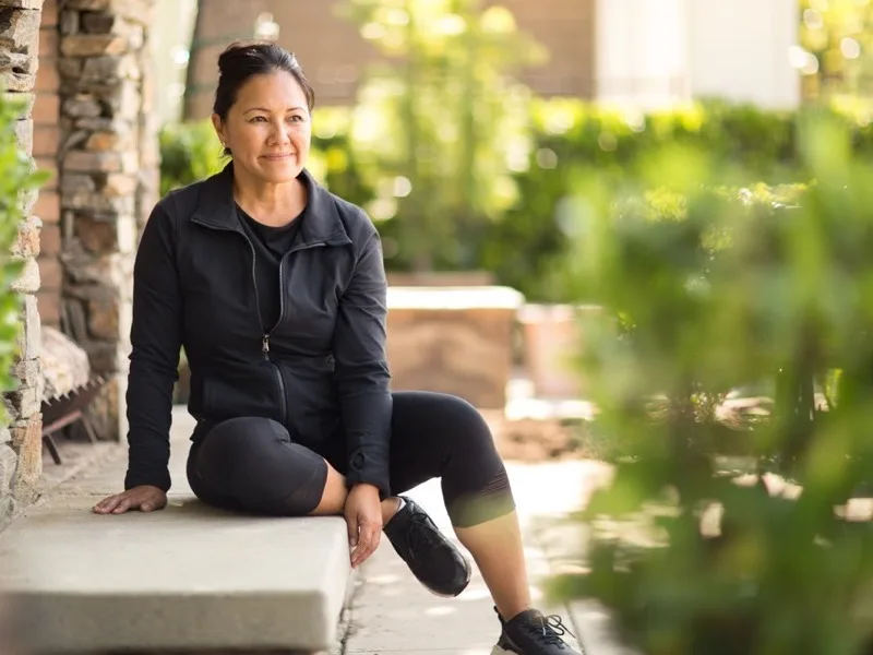 Middle-aged woman in black athletic wear sitting on a stone bench outdoors, smiling.