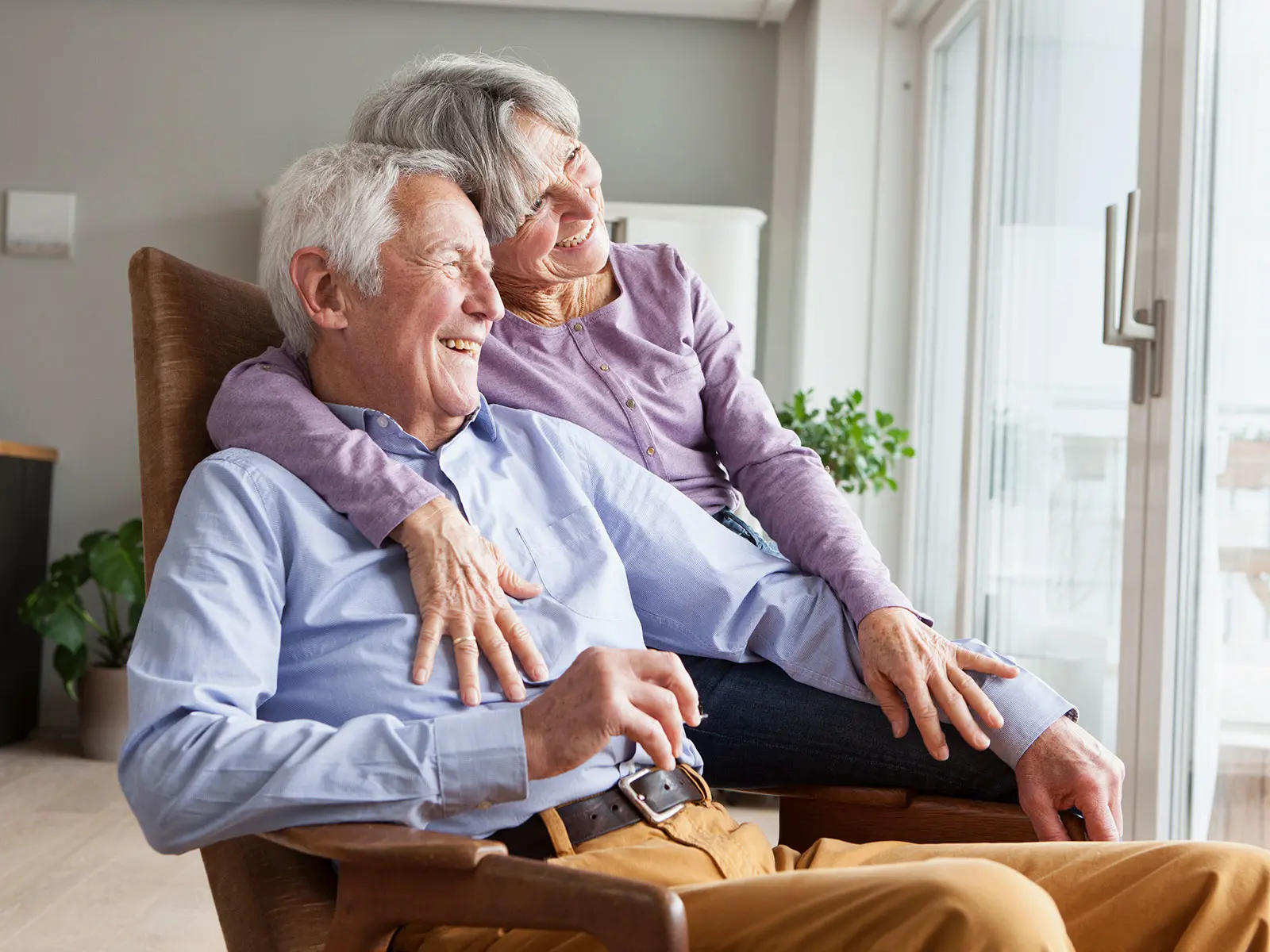 An elderly couple are sitting together on a chair. They are looking outside and seem happy.