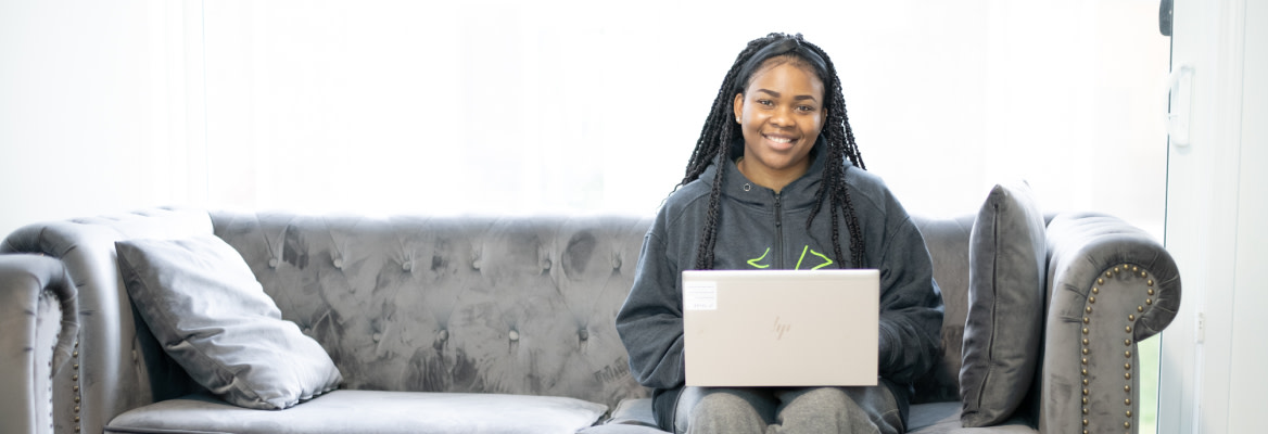A smiling Black woman sits on sofa using a laptop
